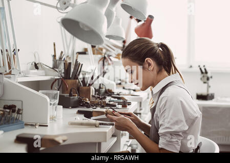 Toute la journée de travail. Vue latérale du bijoutier jeune femme assise à son atelier de joaillerie et tenant en mains les outils nécessaires pour créer des bijoux accessoires. Bijoux Banque D'Images