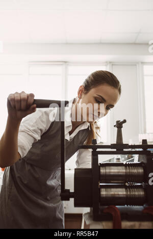 Méthode de travail manuel. Portrait of young female goldsmith ouvrant metal sur le matériel roulant de la machine en atelier. Processus de fabrication de bijoux. L'entreprise. Bijoux wor Banque D'Images