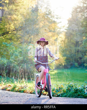 Mode femme Slim blonde attrayant dans les verres, robe courte et rose hat équitation dame location le long de l'allée pavée sur beau parc vert et arbres d'or éclairées par l'arrière-plan lumineux soleil d'automne. Banque D'Images