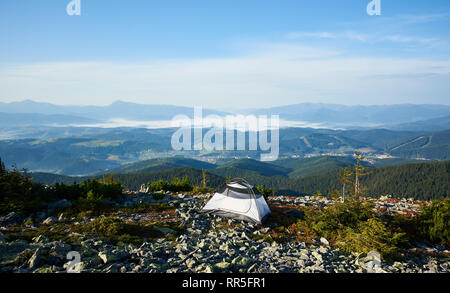 Camping d'été sur le sommet de la montagne avec la vallée brumeuse, de pins et de montagnes lointaines sur l'arrière-plan. Tourisme blanc tente sur rocky Hill dans la matinée. Tourisme et de la beauté de la nature concept Banque D'Images