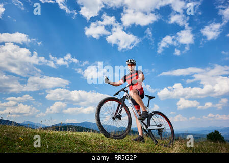 Smiling cycliste professionnelle dans les vêtements de sport et le casque de vélo cross-country avec commandes sur le haut de la colline, showing Thumbs up, contre le ciel bleu avec des nuages sur l'été journée ensoleillée. Outdoor sport concept Banque D'Images