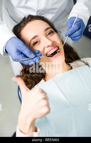 Portrait de dentiste dans des gants en latex examinant cheerful woman en accolades avec bouche ouverte showing thumb up Banque D'Images