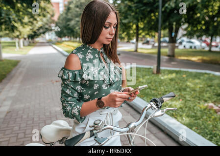 Belle fille avec le téléphone à regarder des vidéos sur Internet, réseaux sociaux, en ligne, bicyclette en été dans la ville. Une femme à l'écoute de la musique sur Banque D'Images