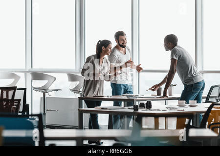 Trois hommes d'affaires dans les vêtements de la conversation tout en comité permanent par table en bois clair et moderne, à l'office. Ils sont souriants et regarder tout en Banque D'Images