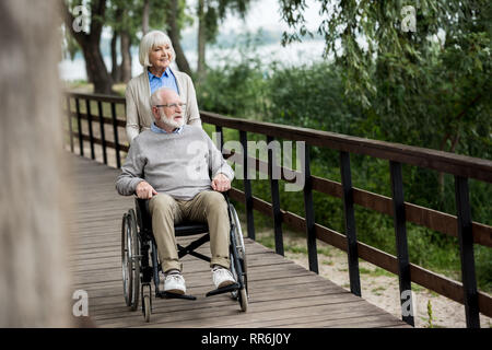 Smiling senior woman avec son mari en fauteuil roulant alors que walking in park Banque D'Images