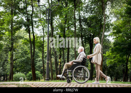 Hauts femme transportant mari en fauteuil roulant alors que walking in park Banque D'Images