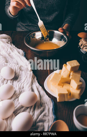 Woman cooking la pâte pour gâteau. Les mains de femelle fouettant œufs avec le sucre dans un bol à côté d'oeufs et les dés de beurre sur table en bois brun. Banque D'Images