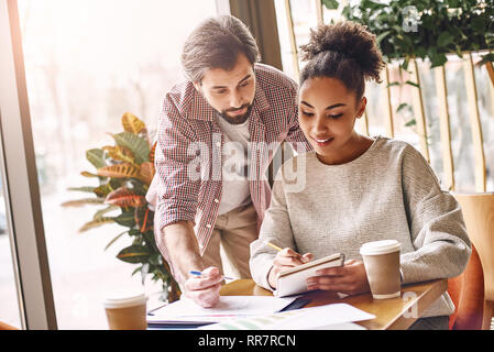 Deux collègues discutent de nouvelles idées gaies dans office . Homme barbu est en train de parler à son collègue, alors qu'elle est la prise de notes. Une tasse de café, les cartes sont sur le bureau. Style décontracté. Concept de la réussite Banque D'Images