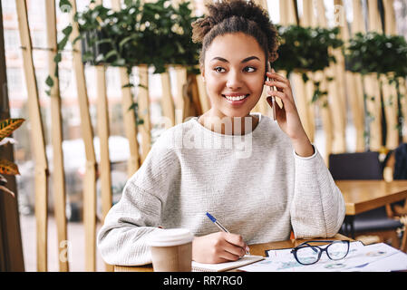 Portrait of happy young woman sitting at her desk talking on mobile phone. Smiling business woman talking avec client sur smart phone in office et à côté. Woman working at desk in office moderne. Ses lunettes et le café sont sur la table Banque D'Images