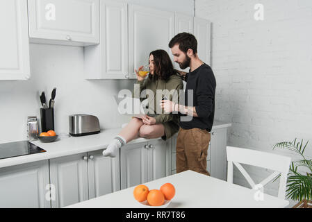 Man hugging woman et boire le jus d'orange au petit-déjeuner dans la cuisine Banque D'Images