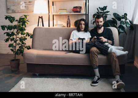 Belle jeune couple sitting on couch avec joysticks et playing video game in living room with copy space Banque D'Images