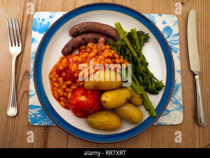 Un Yorkshire repas de saucisses, haricots, tomates grillées, pommes de terre nouvelles et tenderstem le brocoli sur une plaque légèrement bleu sur une table en bois Banque D'Images