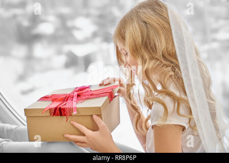 Belle fille avec de longs cheveux bouclés, holding gift avec surprise. Heureux enfant ouvrant grande plaisance fort avec red bow. Adorable, joli adolescent posant près de Banque D'Images