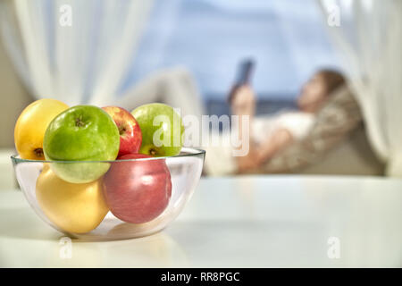 Bol en verre de rouge, jaune, vert pommes fraîches placées sur le tableau blanc dans la cuisine. Girl de bas de veuve ou canapé près de fenêtre dans appartement confortable. Banque D'Images