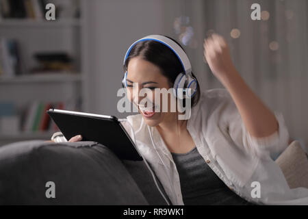 Excited woman écoute et regarde media sur tablet assis sur un canapé dans la nuit à la maison Banque D'Images