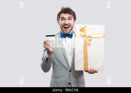 Portrait de jeunes adultes en homme satisfait manteau gris et bleu noeud papillon debout et tenant présent avec bow jaune banque et carte de crédit, à la recherche de s Banque D'Images