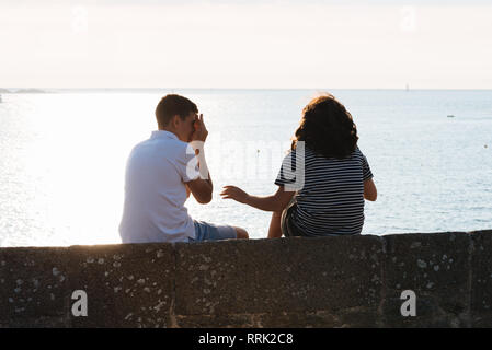 Vue arrière du couple de jouer alors qu'il était assis sur le mur pendant le coucher du soleil contre l'ocean Banque D'Images