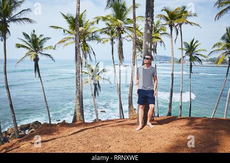 Jeune homme avec des lunettes à vous détendre sous les palmiers contre la mer. Littoral au Sri Lanka. Banque D'Images