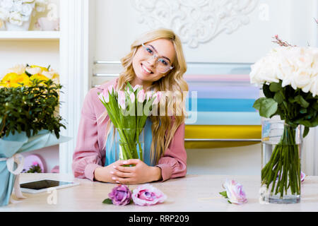 Belle female florist looking at camera et holding vase avec des tulipes roses in flower shop Banque D'Images