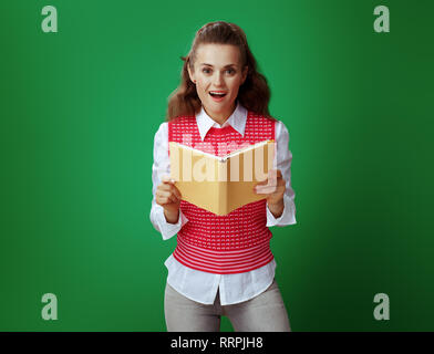 Surpris en étudiant moderne jeans gris et rose T-shirt holding ouvert livre jaune contre tableau fond vert. Banque D'Images