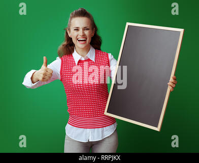 Jeune étudiant en jeans gris et rose T-shirt avec tableau noir showing Thumbs up isolé sur fond vert. Banque D'Images