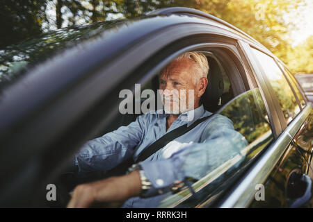 Senior man smiling alors qu'il était assis seul dans sa voiture le long d'une route bordée d'arbres dans le pays Banque D'Images