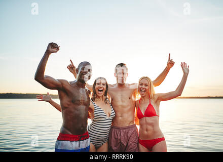 Groupe de rire insouciant young friends standing arm in arm ensemble tout en s'amusant au bord d'un lac en fin d'après-midi Banque D'Images