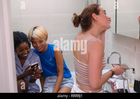 Les jeunes femmes colocataires à l'aide de smart phone et se préparer, appliquant le maquillage dans l'appartement salle de bains Banque D'Images
