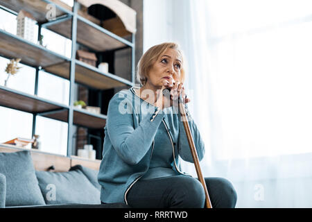 Sad senior woman sitting on sofa holding et bâton de marche à la maison avec copie espace Banque D'Images