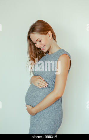 Portrait of smiling white Caucasian blonde femme enceinte robe gris de toucher son ventre. Belle jeune fille attend sur fond clair. Health Banque D'Images