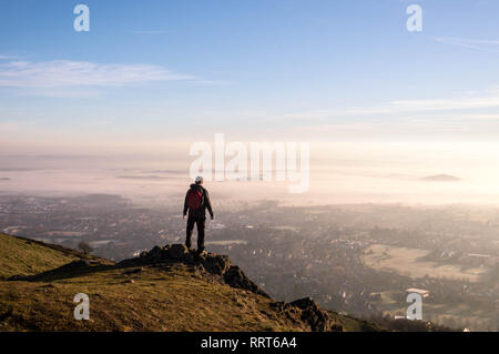 Un randonneur solitaire debout sur un rocher donnant sur un hivers brumeux lever du soleil, Worcestershire Beacon, collines de Malvern, Royaume-Uni. Banque D'Images