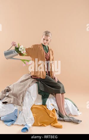 Blonde woman sitting on pile de vêtements et holding watering can avec des fleurs sur fond beige, de l'environnement concept d'économie Banque D'Images