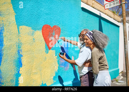 Happy senior couple painting heart-shape murale sur mur ensoleillé Banque D'Images