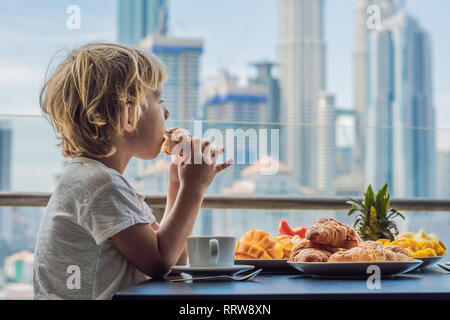 Le garçon est de prendre le petit déjeuner sur le balcon. Table de petit-déjeuner avec café et fruits pain croisant sur un balcon dans le contexte de la grande ville Banque D'Images