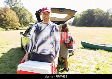 Portrait smiling man carrying camping glacière, déchargement voiture à field Banque D'Images