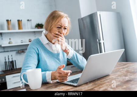 Contrarié senior woman couvrant la bouche avec la main, à l'aide d'ordinateur portable et avoir video chat dans la cuisine Banque D'Images
