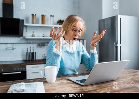 Surpris senior woman gesturing avec les mains, à l'aide d'ordinateur portable et avoir video chat dans la cuisine Banque D'Images