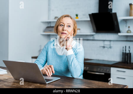 Pensive senior woman sitting at table with laptop and étançonnements chin dans la cuisine Banque D'Images