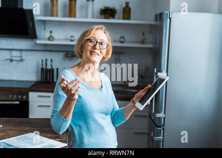 Smiling senior woman in glasses using digital tablet et faisant des gestes avec la main dans la cuisine Banque D'Images