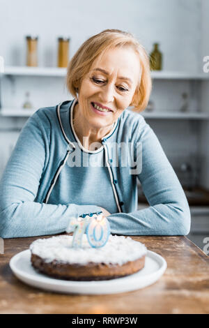 Senior woman sitting at table et à la recherche au gâteau avec '70' au cours d'anniversaire à la maison Banque D'Images