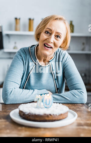 Senior woman sitting at table et à la caméra à gâteau avec '70' au cours d'anniversaire à la maison Banque D'Images