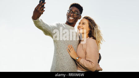 Jeune couple aimant les vacances d'été en prenant un autoportrait avec le smartphone à l'extérieur. Couple Interracial en vacances en tenant contre selfies ciel clair Banque D'Images