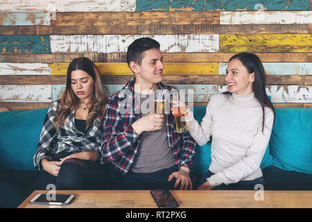 Happy young girl drinking beer avec jeune homme ignorant et rencontrer d'autres jaloux femme triste assise à côté d'eux au bar restaurant rustique. Et j'aime Banque D'Images