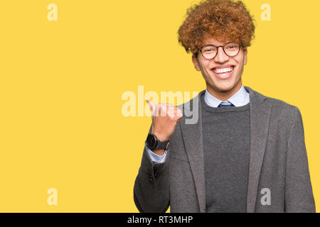 Beau jeune homme d'affaires avec afro portant des lunettes avec sourire heureux face à la recherche et en pointant sur le côté avec le pouce vers le haut. Banque D'Images