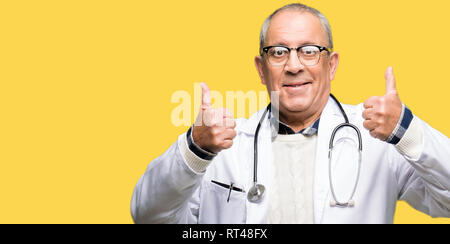 Beau médecin senior homme portant un manteau médical signe de réussite faisant geste positif avec la main, les pouces jusqu'en souriant et heureux. Regardant la caméra avec Banque D'Images