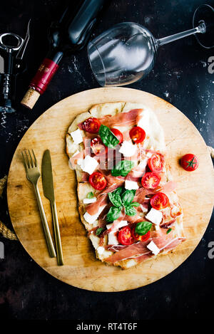 Pain plat fait maison italienne avec tomate, fromage, jambon, feta et basilic, sur une planche à découper ronde sur un tableau noir, télévision coucha avec le couteau et le vin rouge Banque D'Images