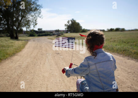 Fille avec drapeau américain équitation vélo sur chemin dans paysage à distance Banque D'Images