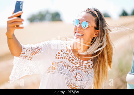 Portrait de jeune femme blonde portant des lunettes de soleil miroir en tenant avec smartphone selfies Banque D'Images