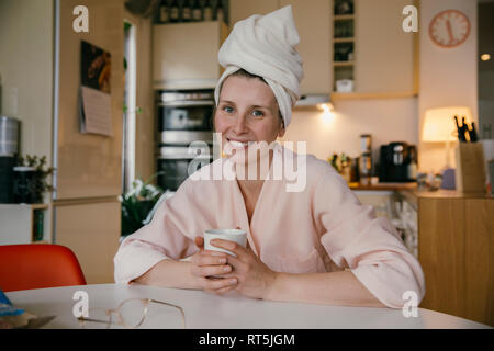 Portrait of smiling woman wearing towel turban assis avec une tasse de café à la table dans la cuisine Banque D'Images