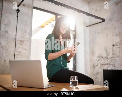 Smiling woman sitting on desk in office d'écouter de la musique Banque D'Images
